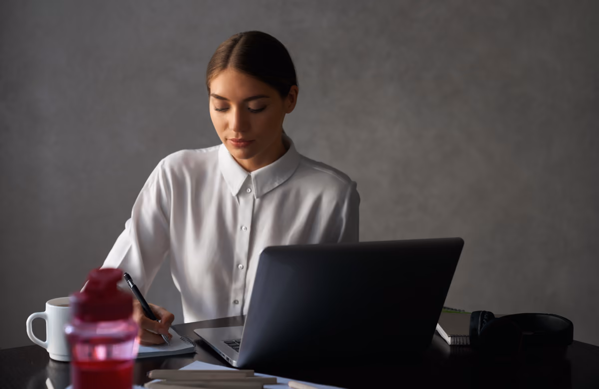 Mujer trabajando con un cuaderno de notas junto a un portátil abierto